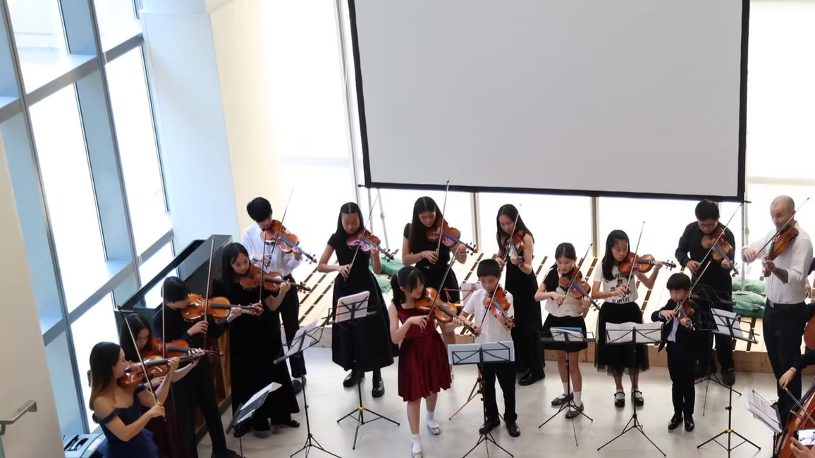 Orchestra of children and adults playing violins during a performance in a bright room with large windows and a blank projection screen.