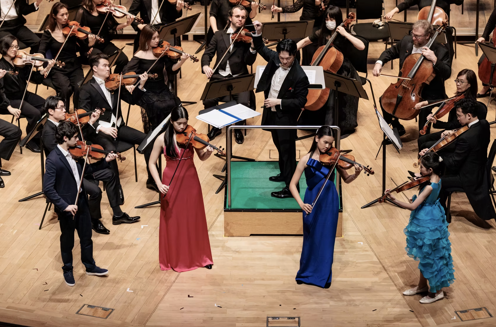 An orchestra performing on stage with four young female soloists in colorful dresses and four male conductors in formal attire.