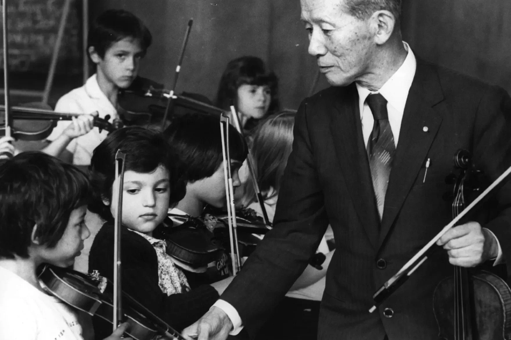 Dr. Shinichi Suzuki conducting a group of children playing string instruments in an orchestra.