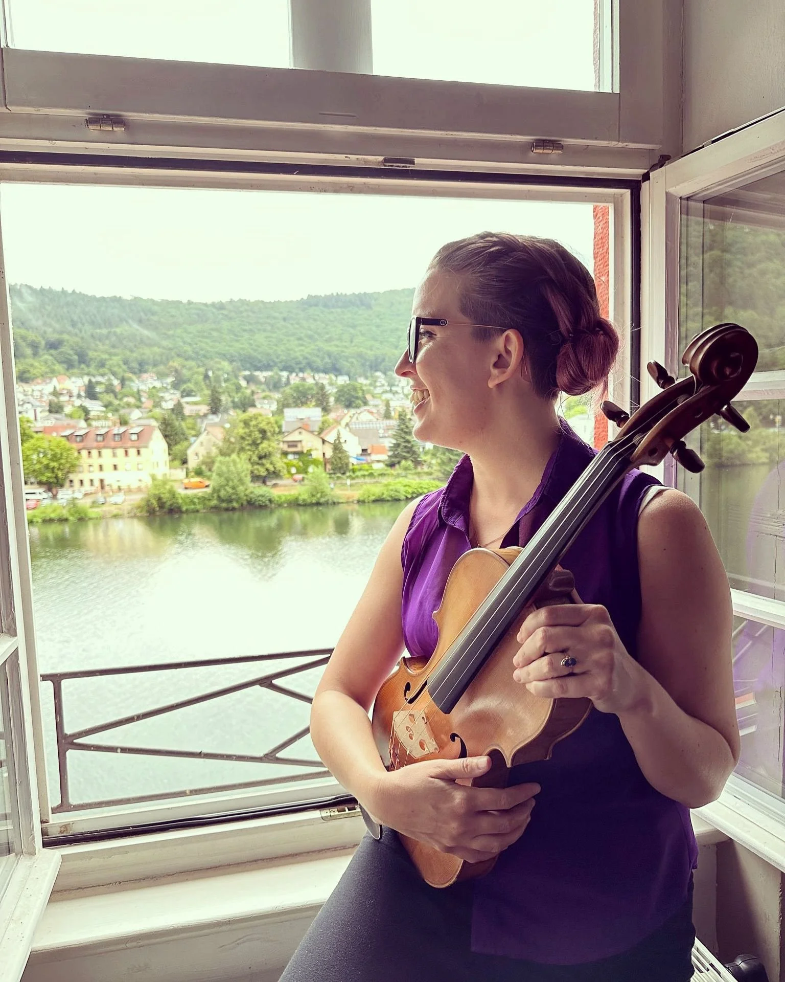 Ms. Monica Johnston: A woman with glasses smiling and playing a violin by a window overlooking a river and a town with houses and green hills.