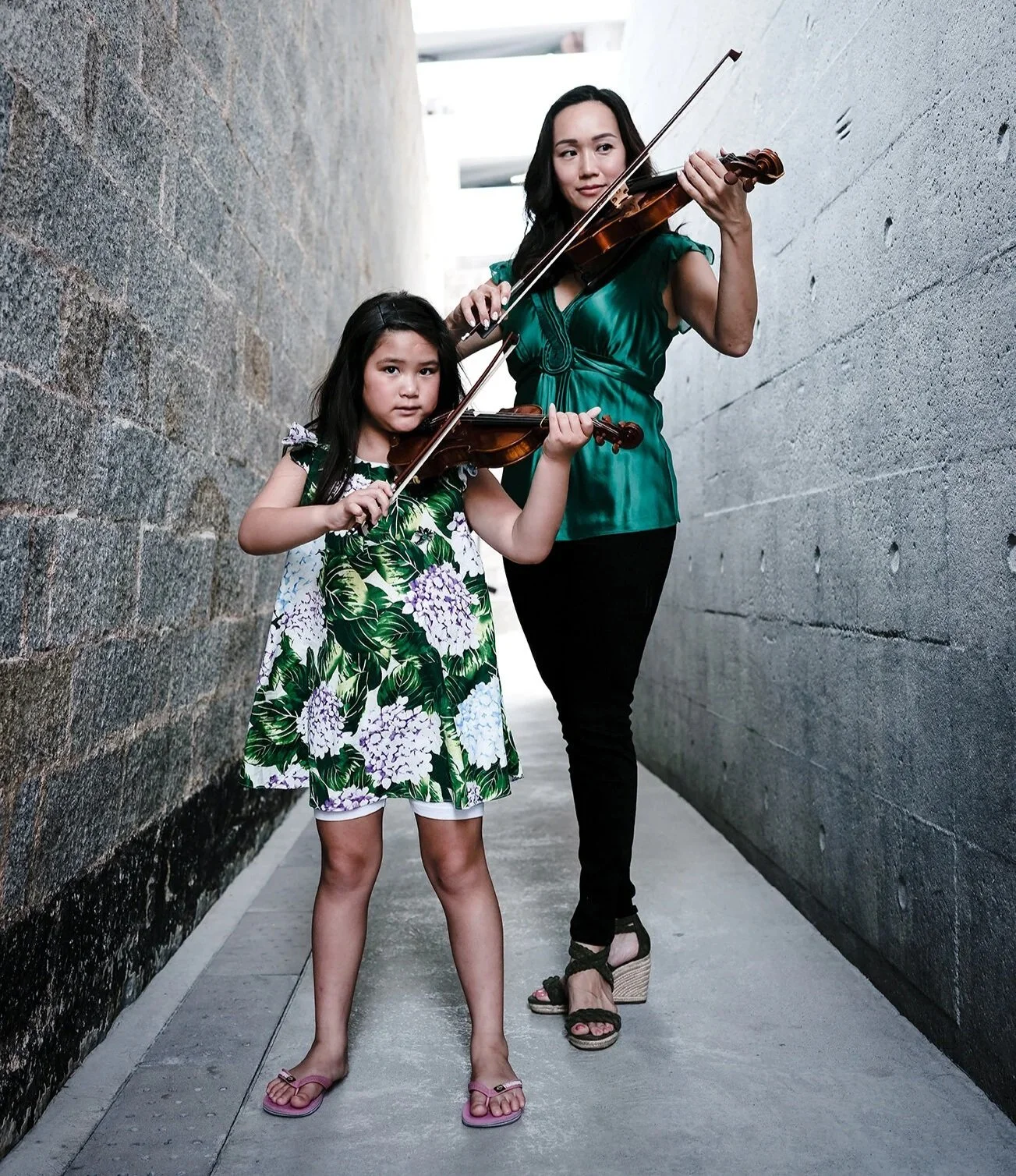 A woman and a young girl are standing in a narrow alley with stone walls, playing violins.