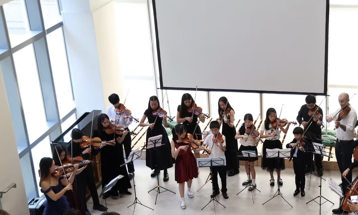 Young Virtousi Program: A group of children and adults playing violins during a performance in a modern indoor space with large windows and a blank projector screen.