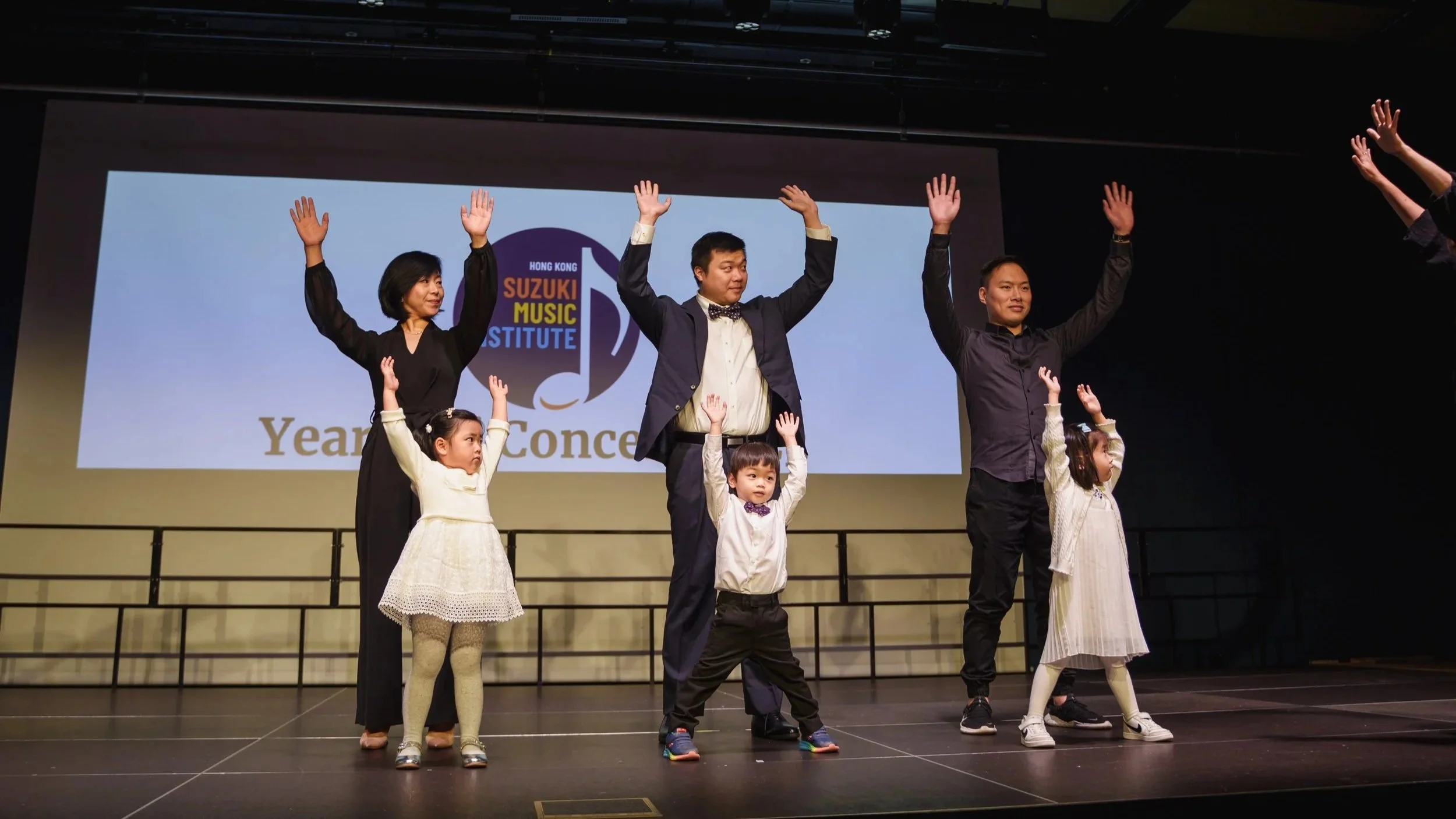 People on stage raising their hands during a performance or presentation at the Hong Kong Suzuki Music Institute, with children in the front row wearing white dresses and suit jackets, and a large screen displaying the institute's logo and event information behind them.