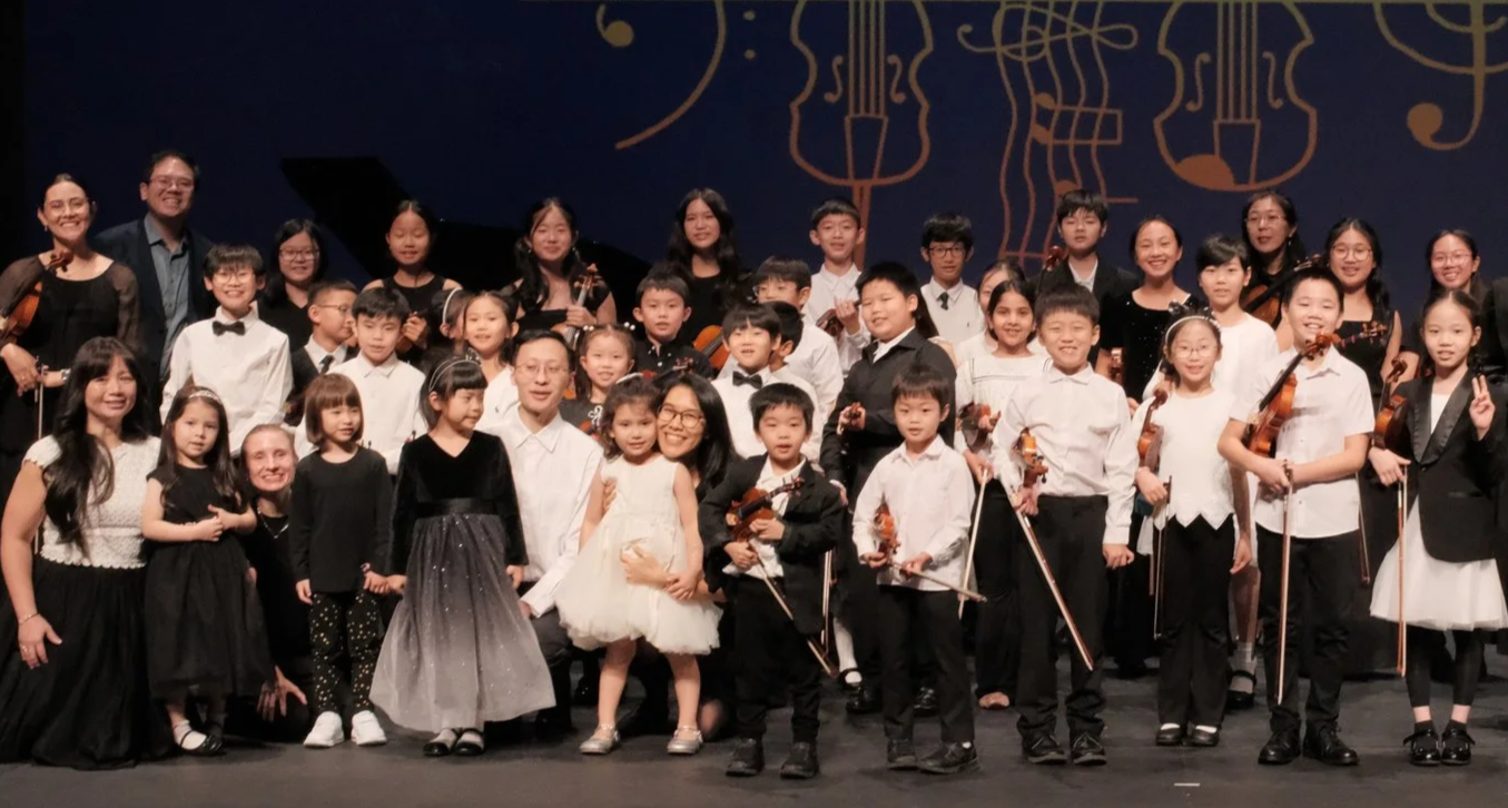 Group of children and adults on stage after a musical performance, some holding violins, surrounded by a musical note-themed backdrop.