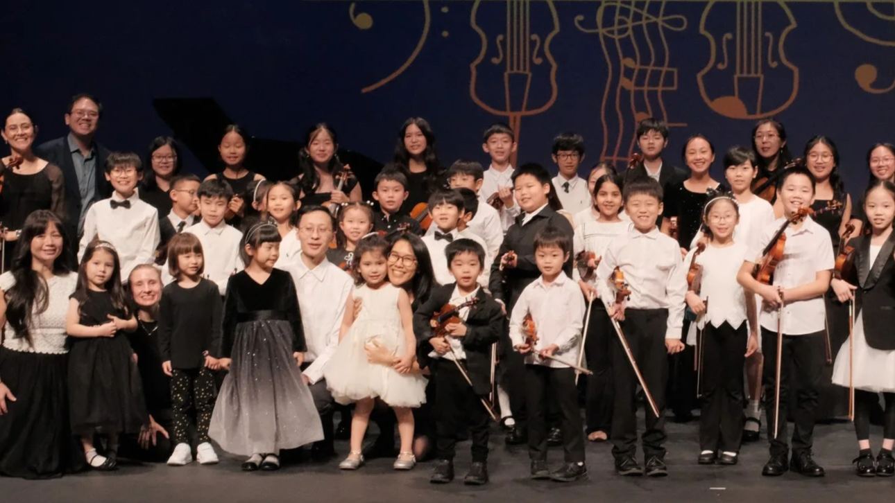 Group of children and adults on stage after a musical performance, some holding violins.