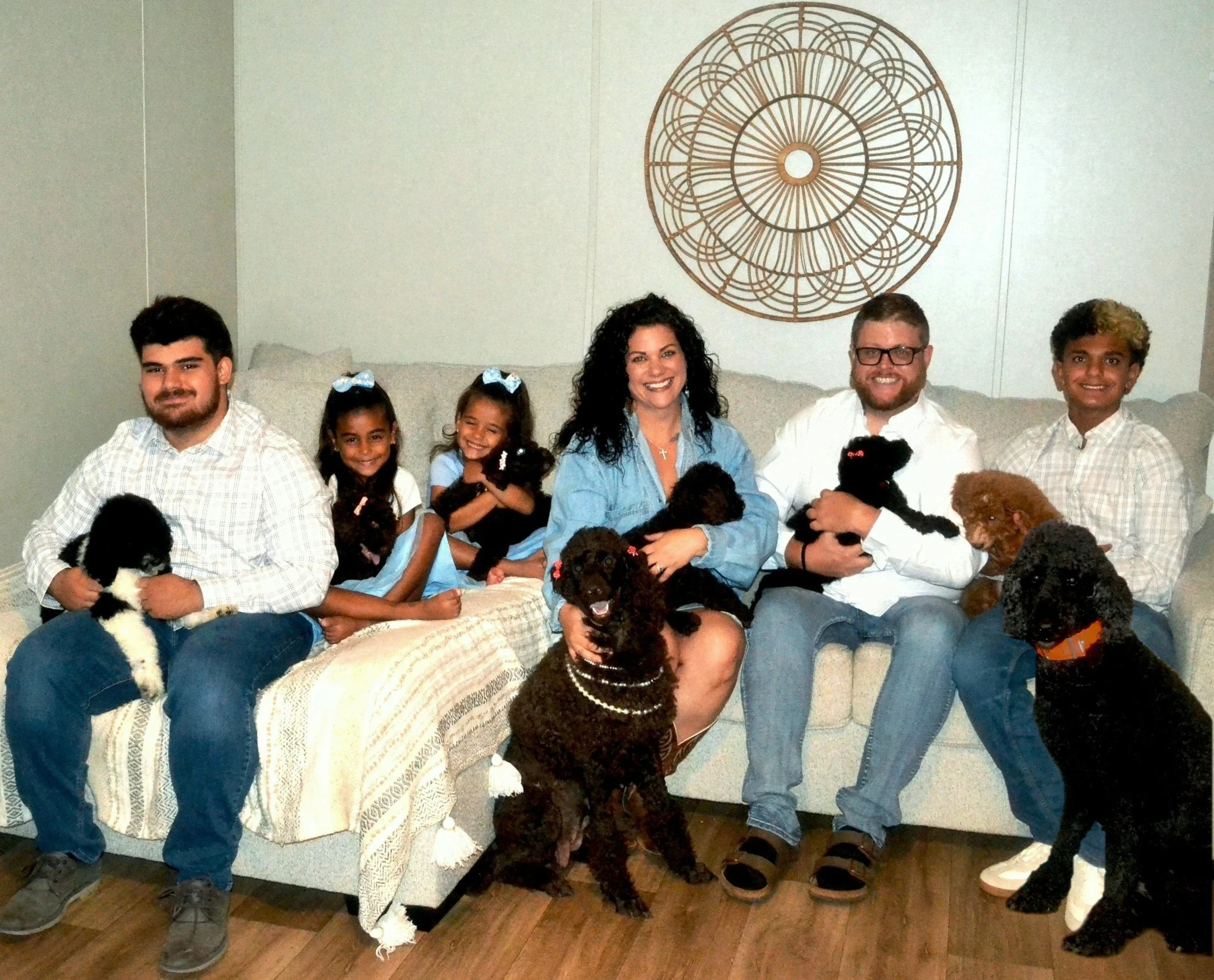 Family of six with six poodle puppies and two adult poodles sitting on an L-shaped couch.