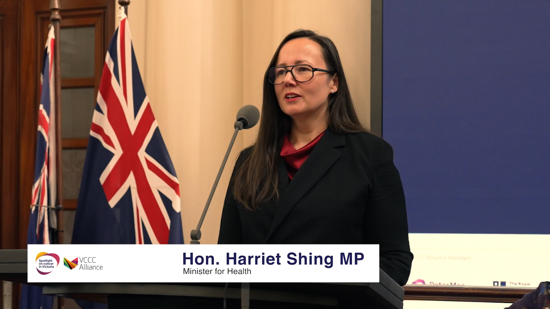 A woman with long dark hair, glasses, and a black blazer giving a speech at a podium with a microphone. Behind her are flags of Australia and another country. A banner at the bottom identifies her as Hon. Harriet Shing MP, Minister for Health, during a formal event.