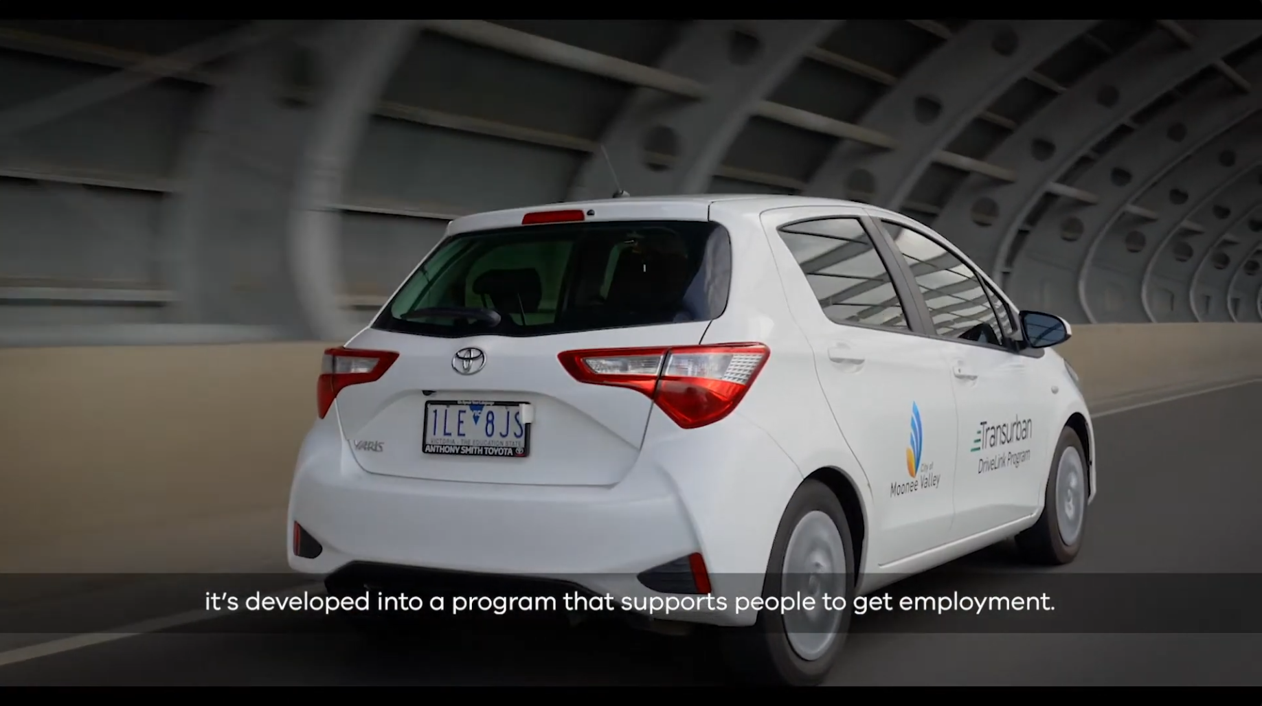 White Toyota Yaris car driving inside a tunnel with a logo and text on the side, and a license plate reading 1LEV8JS.