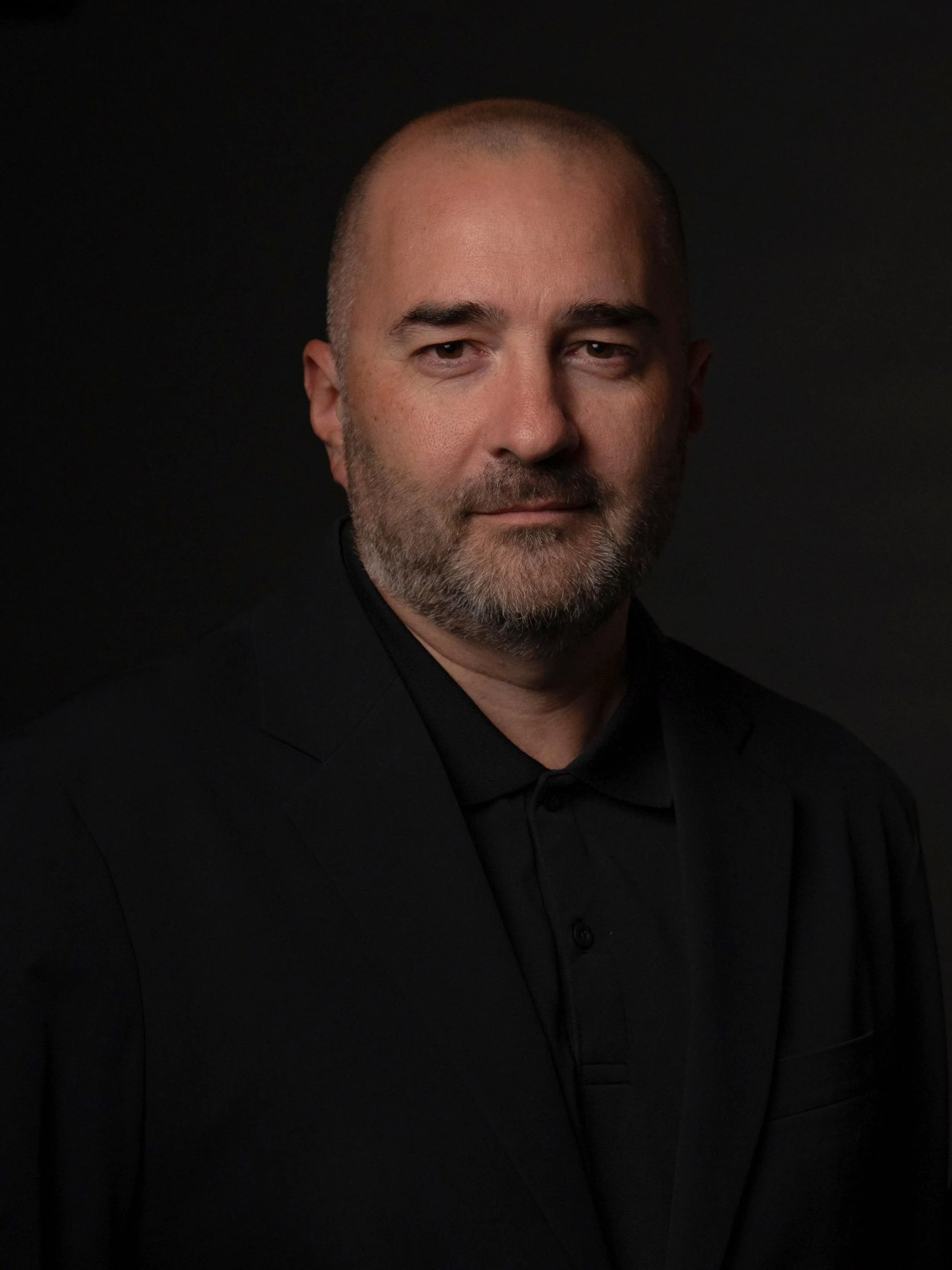 A man with a shaved head and beard wearing a black suit, black shirt, and black tie, standing against a dark background.
