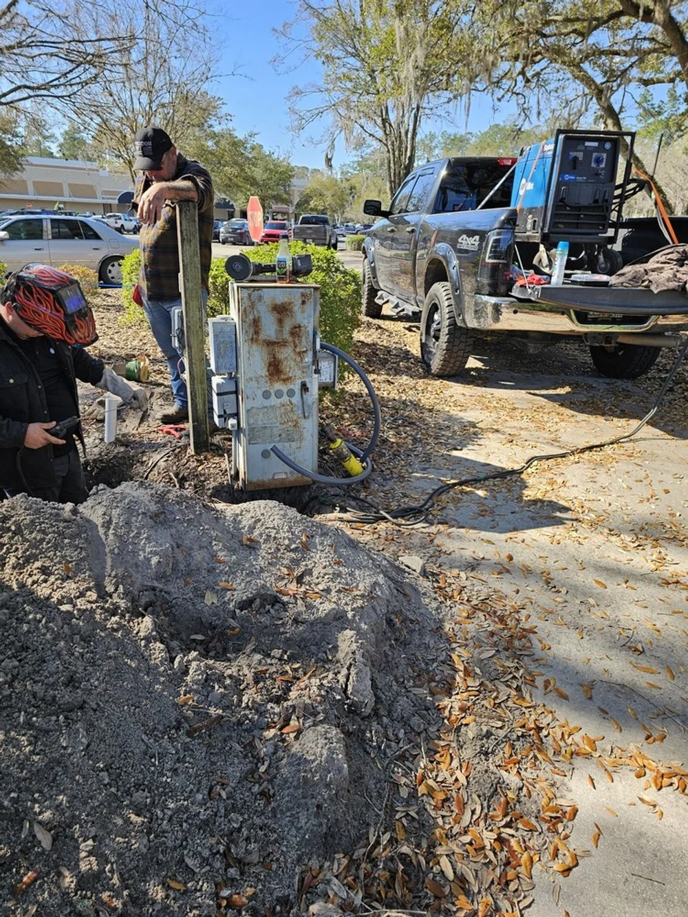 Workers performing underground electrical work near a parking lot, with one person in a helmet and another in a baseball cap, a pickup truck with equipment, and a pile of dirt and leaves on the ground.