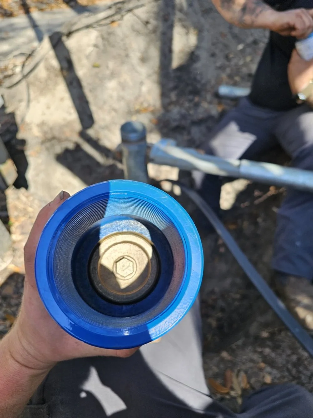 A person holding a blue and clear cylindrical object, possibly a pipe or container, while outdoors on rocky ground with another person seated nearby.