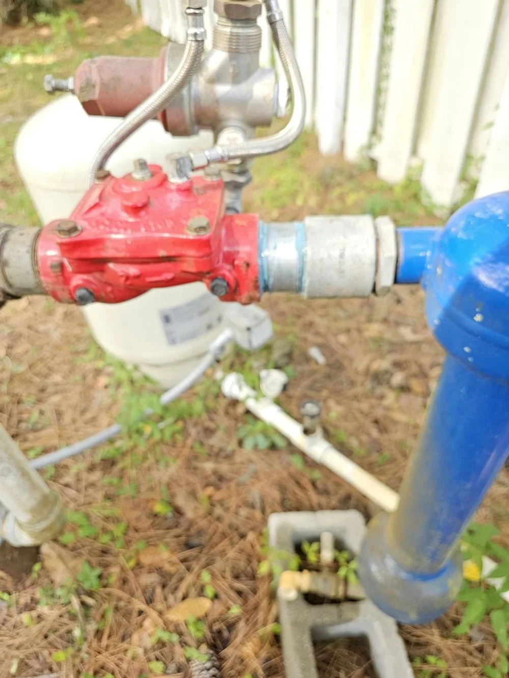 Close-up of outdoor plumbing with a red valve, metal fittings, and blue pipe, with a white propane tank and a white fence in the background.