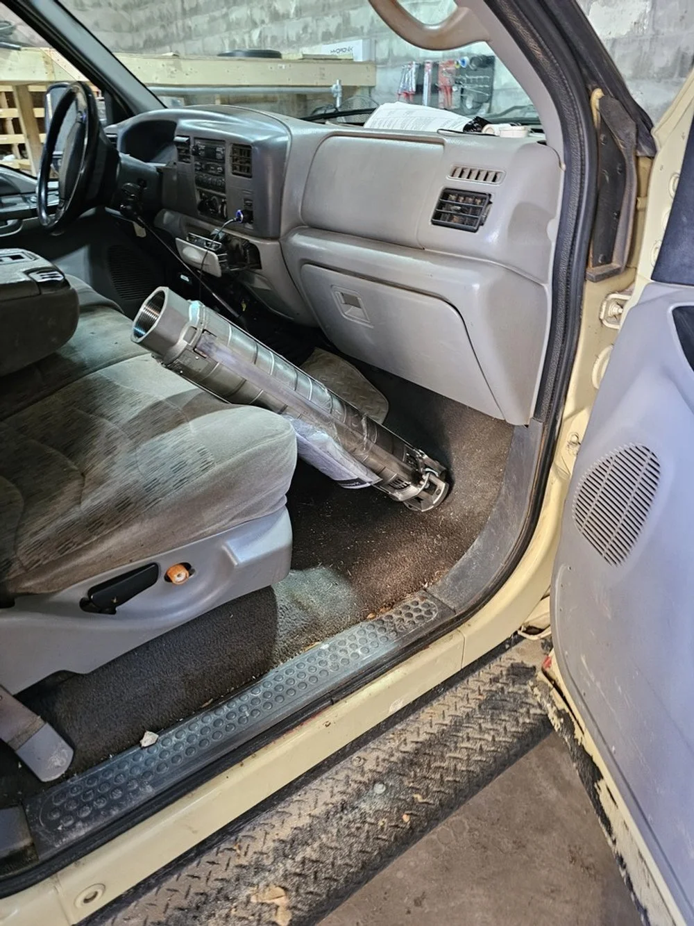 Inside the cab of a work truck, showing the dashboard, steering wheel, and a large tool resembling a rocket launcher resting on the passenger seat.