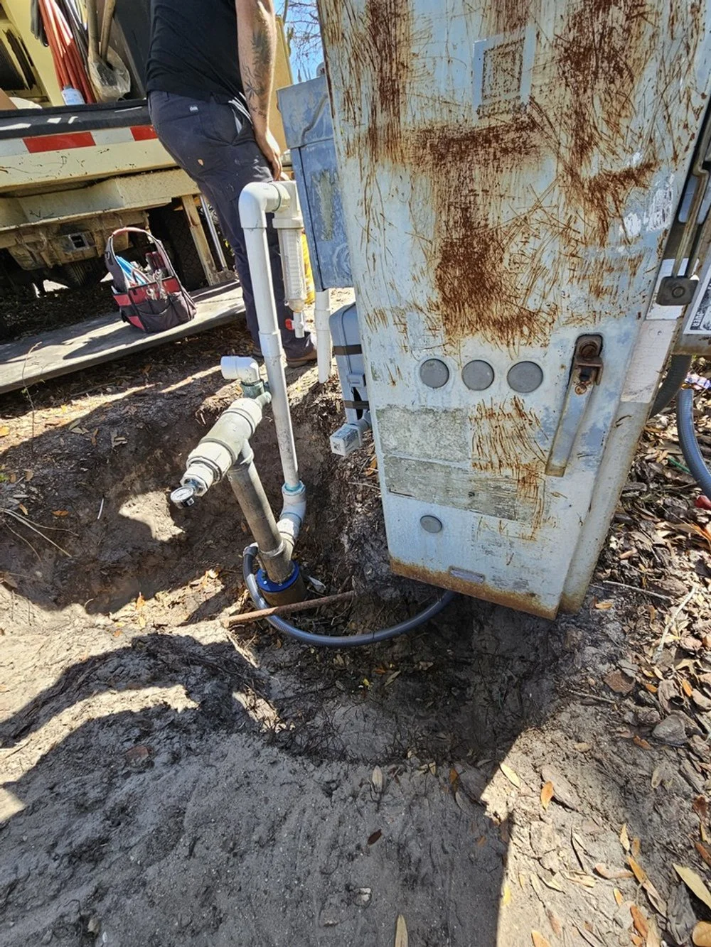 An outdoor utility installation with pipes and wiring connected to a rusty metal box, partially buried in the ground with a person working nearby, a truck in the background, and a toolbox on the ground.