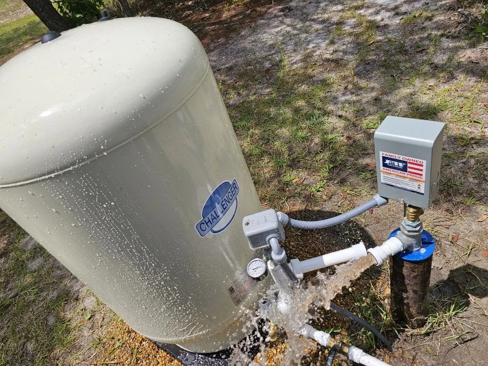A water pump system outdoors with water flowing from a pipe into a large beige water tank labeled 'Challenger' and a control box marked 'Family Owned'.
