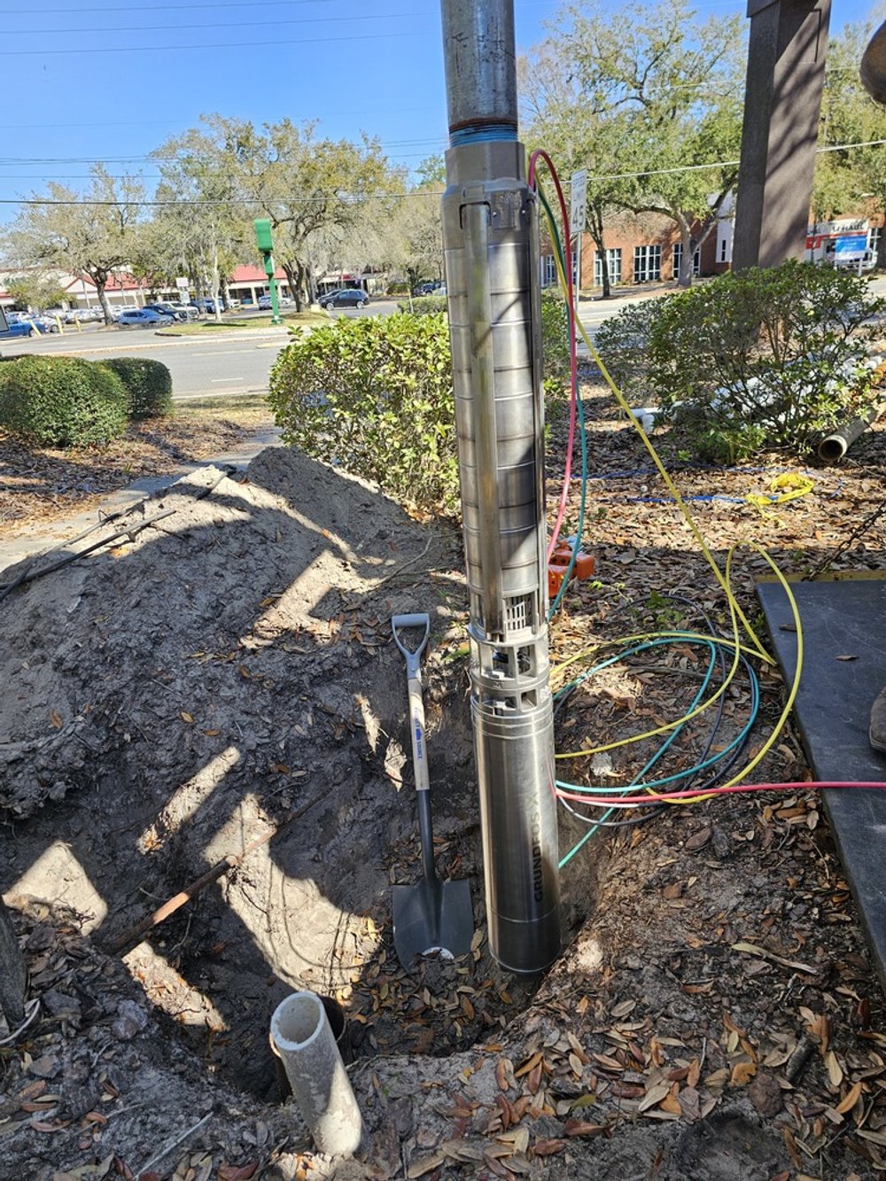 A construction site showing an underground utility vault with connected wires, a shovel in the hole, and a PVC pipe nearby, with bushes, trees, and a street in the background.