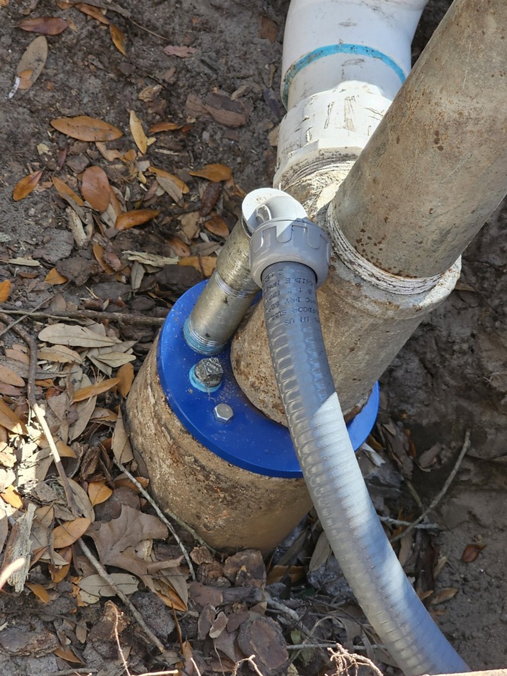 Close-up view of a plumbing pipe connection underground, with a large rusted pipe and a smaller gray pipe, surrounded by dirt and fallen leaves.
