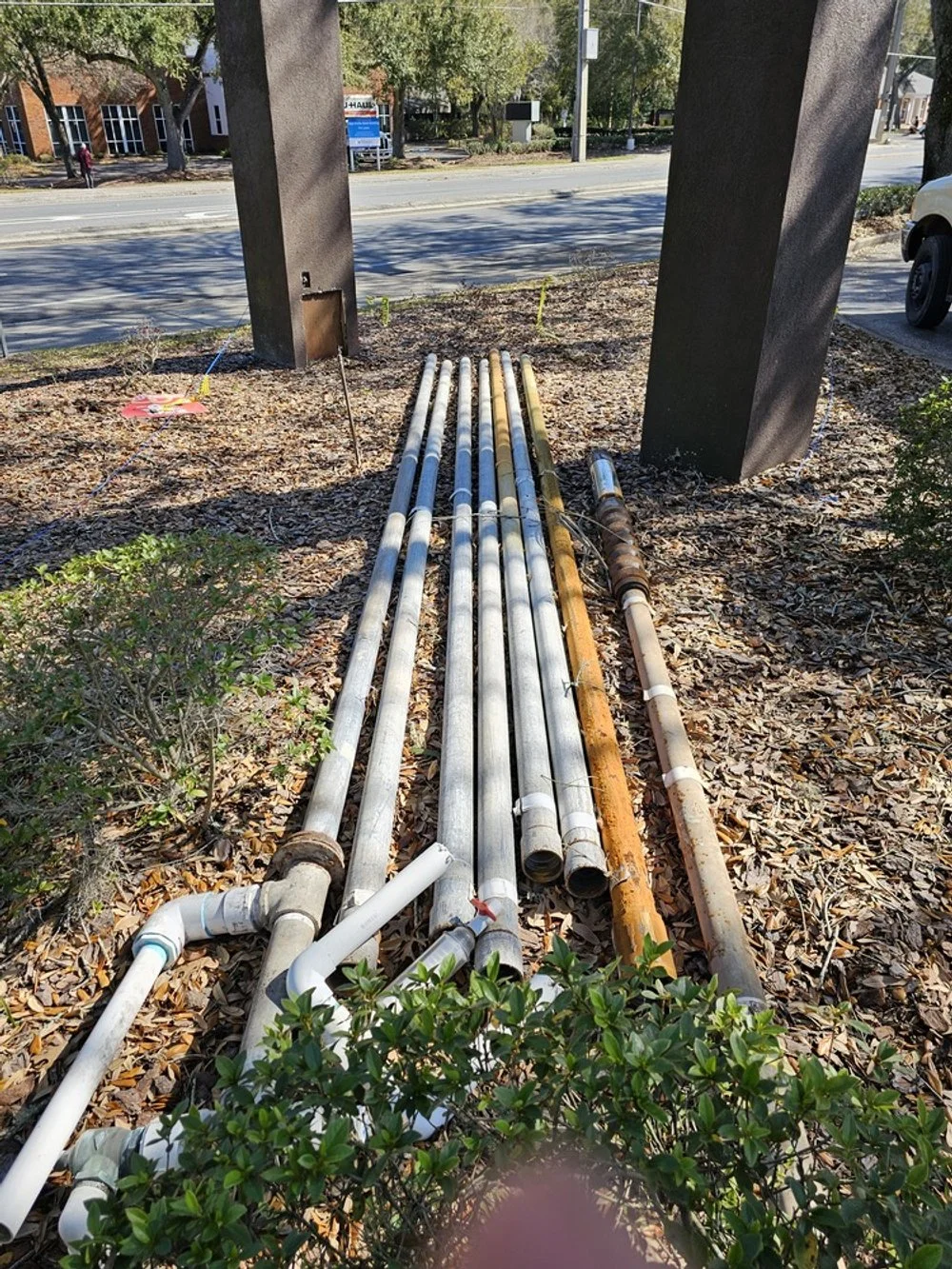 Multiple underground pipes, some white and some rust-colored, are laid out on the ground next to two large black columns, with tools and markers nearby, indicating construction work.