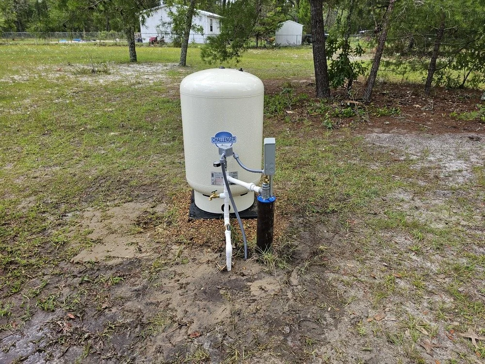 An outdoor irrigation system features a white tank labeled 'Challenger' connected to hoses and pipes, set on a patch of grass and dirt surrounded by trees.