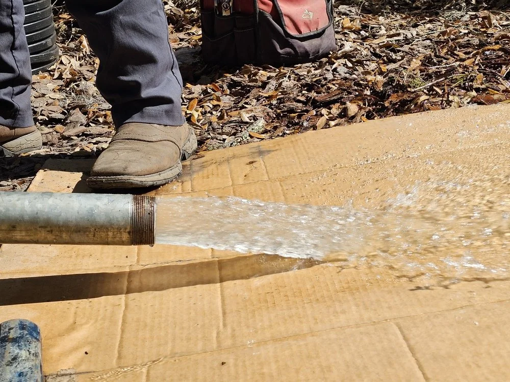 A worker's shoes and pant legs near a pipe that is spraying water onto a piece of cardboard on the ground outdoors.