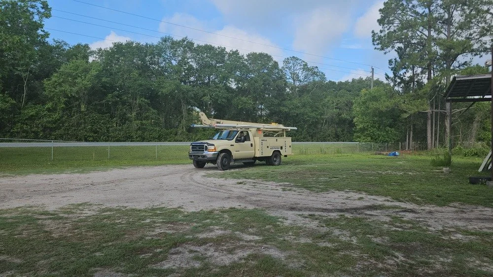 A beige utility truck with a ladder on top, parked on a dirt and grass area near a chain-link fence, with trees in the background and a partly cloudy sky.