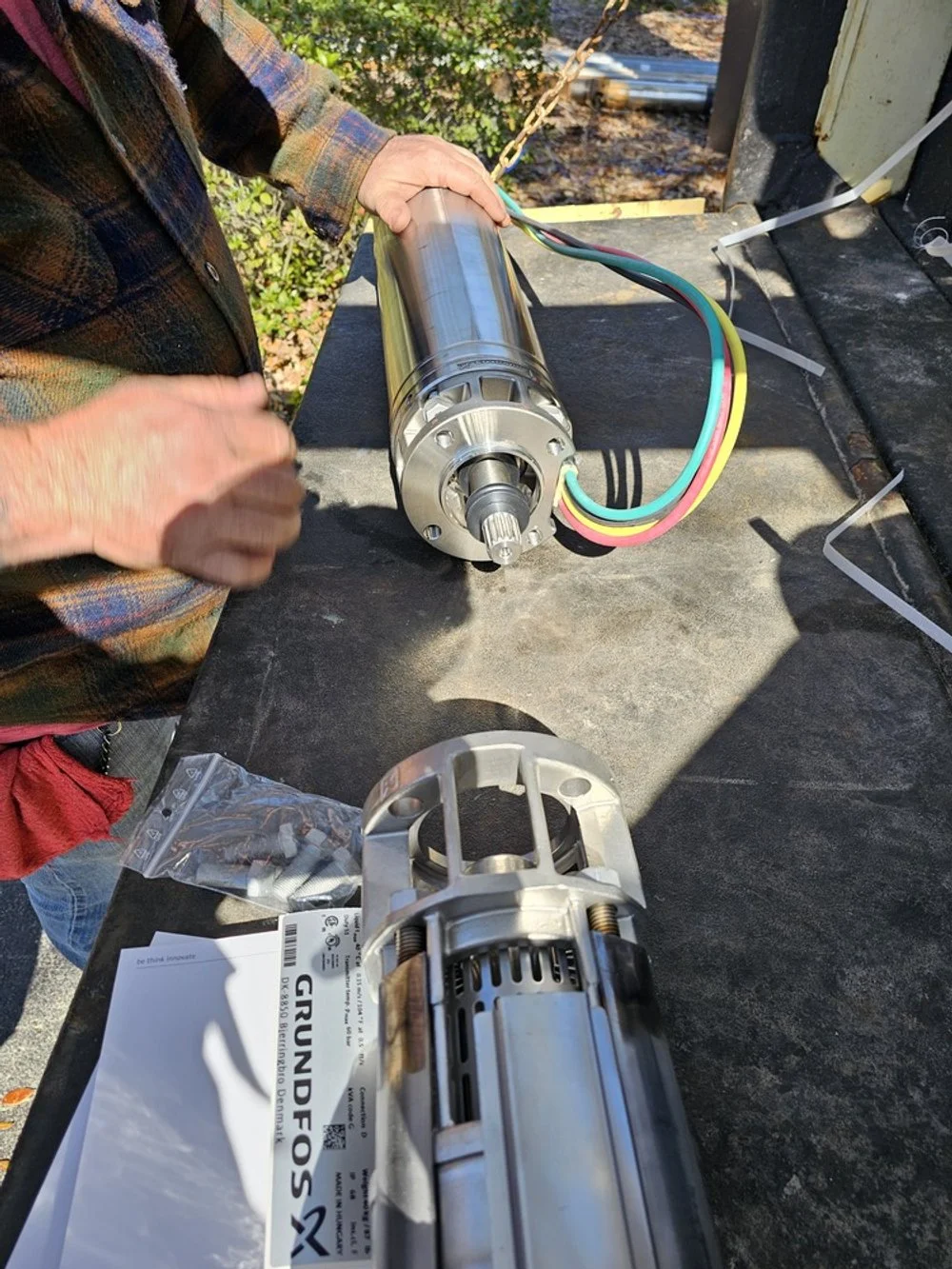 Person working outdoors on a large electric motor or generator, with wires connected to it, placed on a black work surface. Tools and packaging nearby.