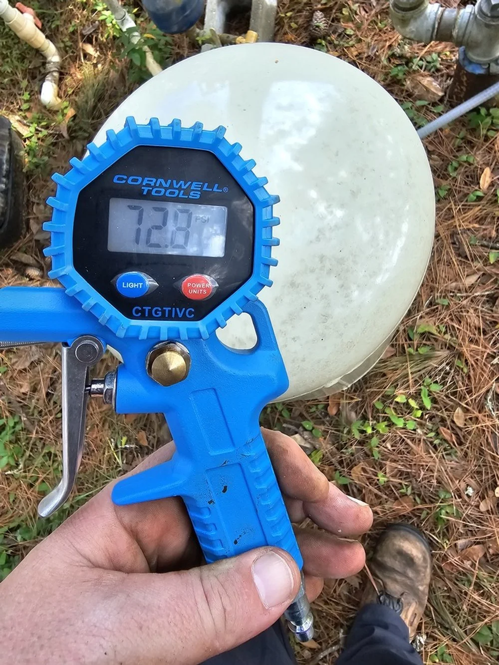 A hand holding a compressor gauge reading 72.8 PSI next to an outdoor propane tank surrounded by pine needles and grass.