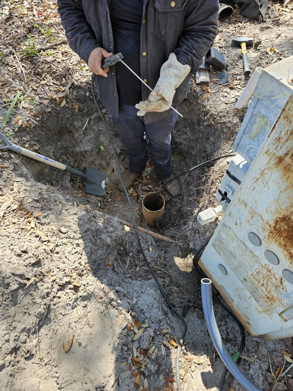 Worker in gloves using a measuring tape in a hole near underground pipes, with tools and equipment nearby.