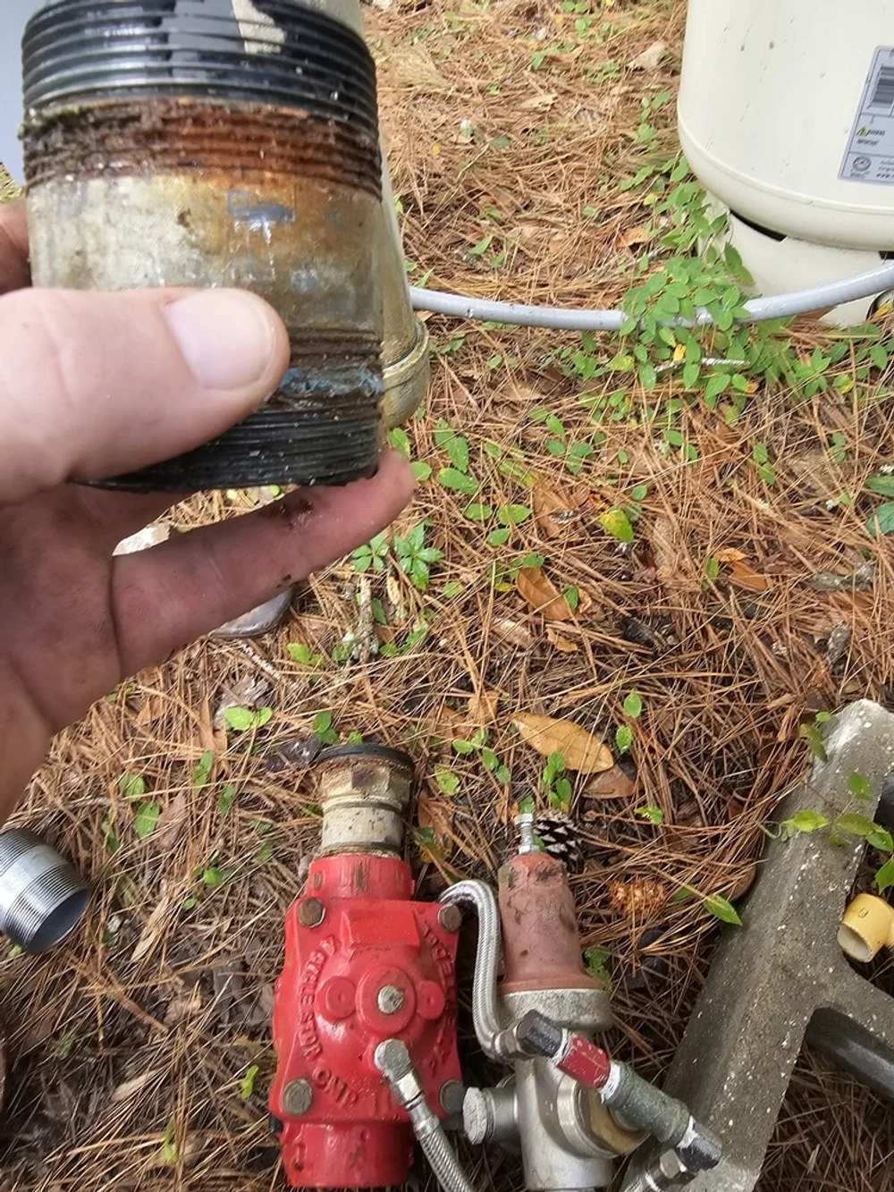Close-up of a hand holding a rusty, black pipe fitting outdoors, with a red valve and plumbing components on the ground nearby and a white water heater in the background.