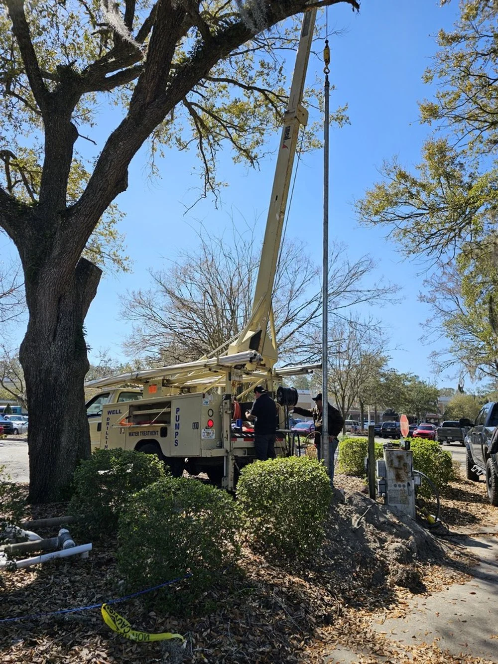 Workers repairing or maintaining a streetlight or utility pole in a parking lot with trees and parked cars in the background.