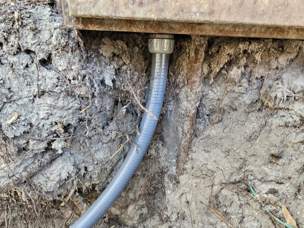 A gray conduit pipe coming out of the ground near a buried structure, with soil and small roots around it.