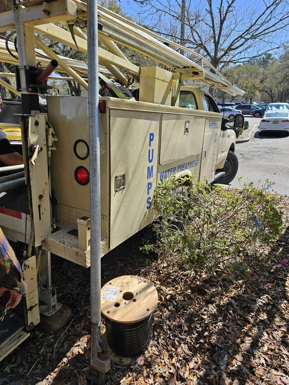 A yellow well drilling truck labeled 'PUMPS' and 'WATER TREATMENT' is parked outdoors among trees and other vehicles. It has an extended ladder on top and equipment attached to the back, with a spool of wire and some bushes in the foreground.