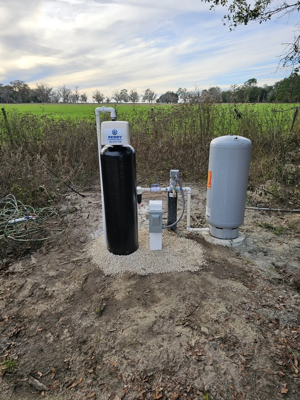 Outdoor well water system with black filtration tank on the left and gray pressure tank on the right, installed on a dirt patch with a rural background of green fields and trees.