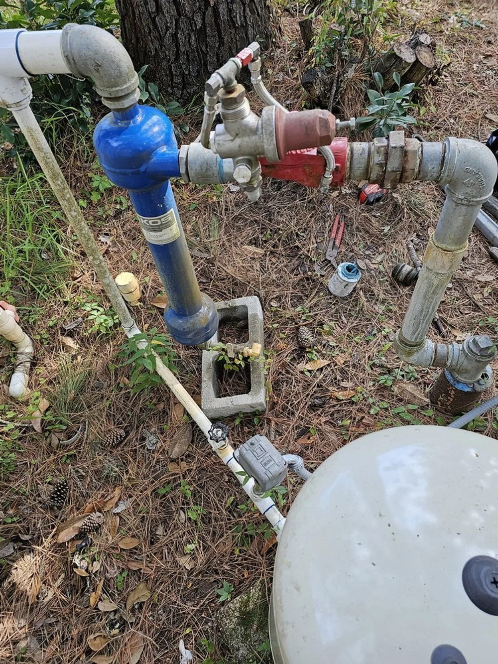 Outdoor plumbing setup with pipes, valves, and a water filter system on a ground covered in pine needles and leaves.