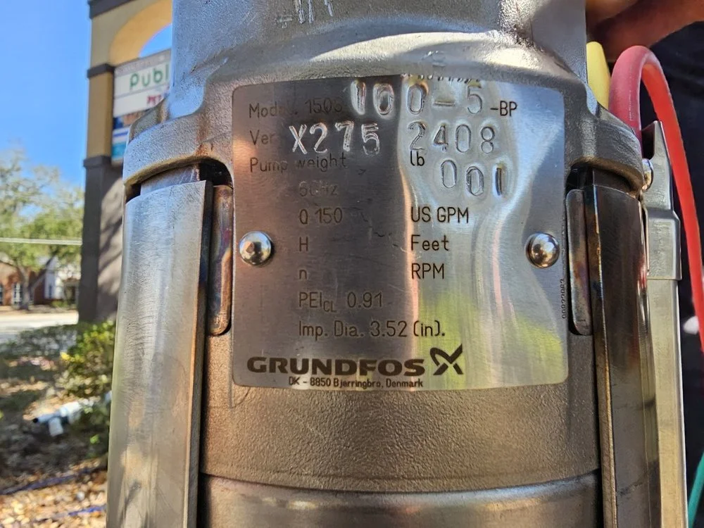 Close-up of a metallic pump or motor with a label displaying model number, serial number, pump weight, and other technical specifications, with outdoor background including a building, blue sky, and some trees.
