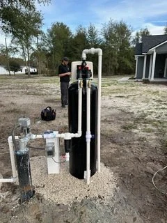 Person standing outdoors next to a large water filtration system connected to white pipes, with houses and trees in the background.