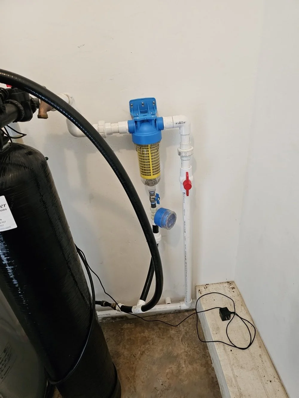 Water filtration system with blue filter housing, white pipes, red valve, and black water tank in a utility room.