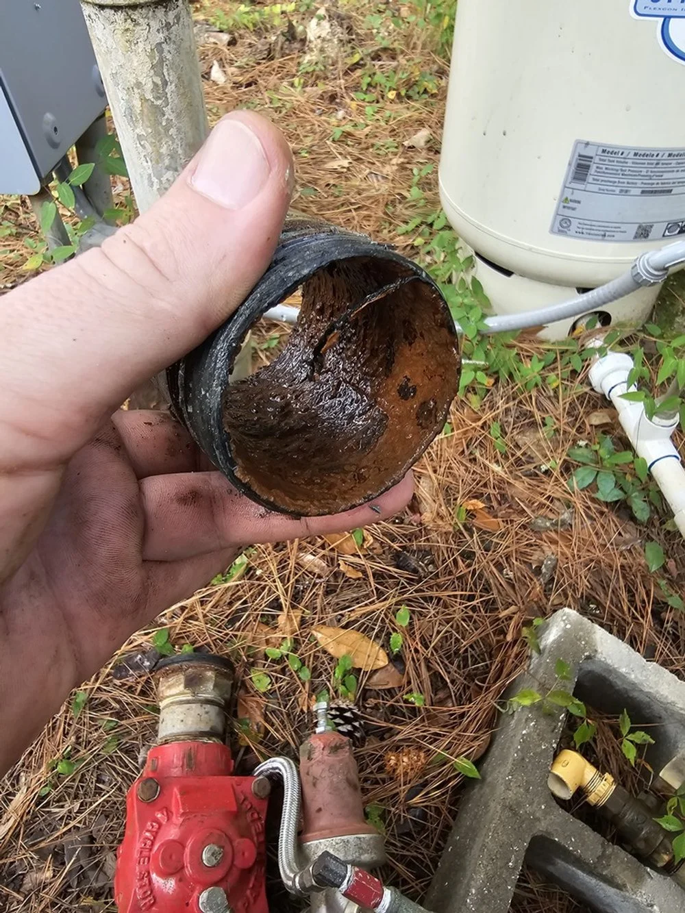Person holding a damaged, rusty pipe or fitting near outdoor plumbing components, including a white water heater and red valve, surrounded by pine needles and small plants.
