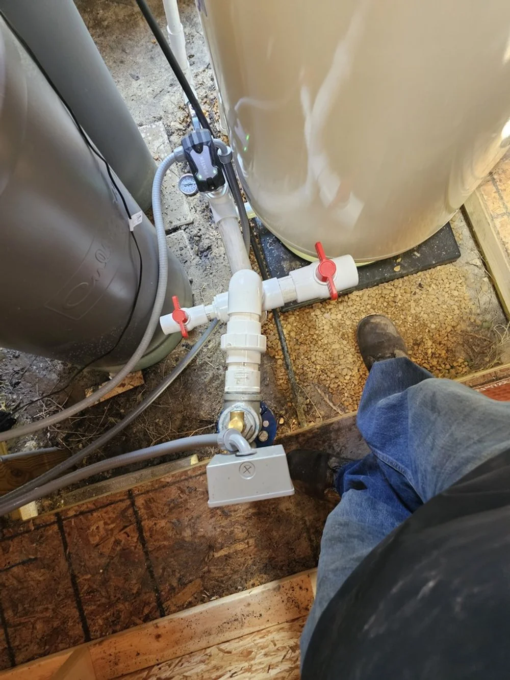 View of a person's lower body standing on a concrete and dirt floor, inspecting or working on plumbing pipes connected to a large tank and a gray water filtration system or pump system in a basement or utility room.