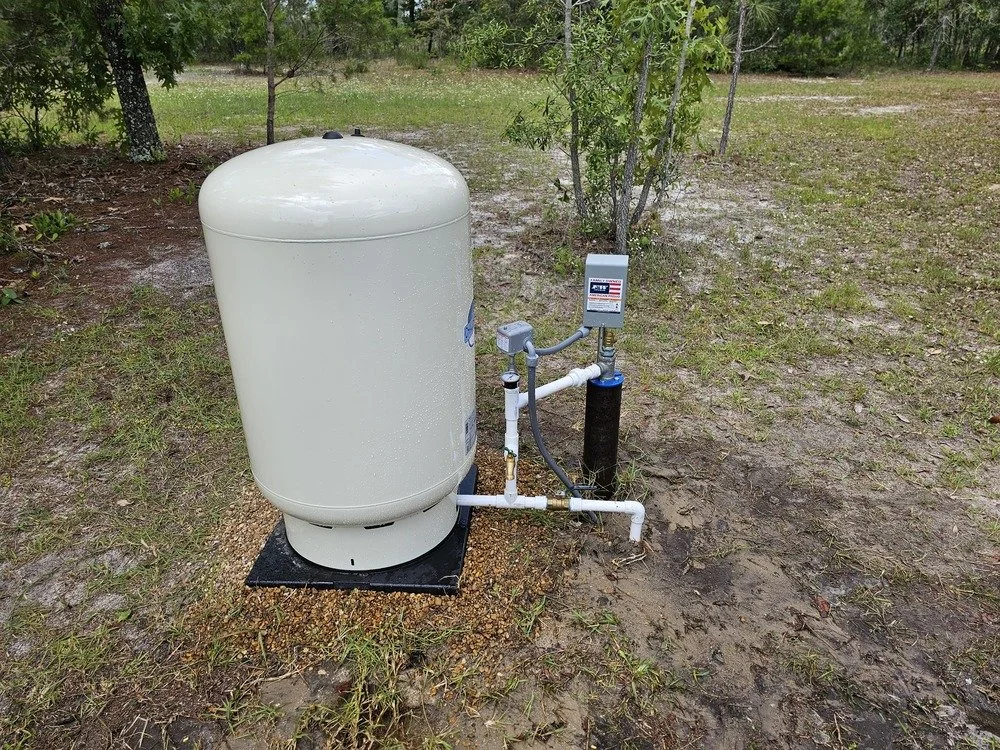Outside well or water pressure tank system with pipes and control box on grassy soil area with trees in background.