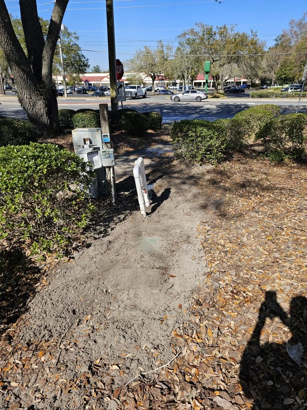 Sidewalk in a park area with underground utility or cable boxes, a meter, and some pipes. There are bushes, fallen leaves, and shade from trees. A busy street with cars and a shopping center can be seen in the background.