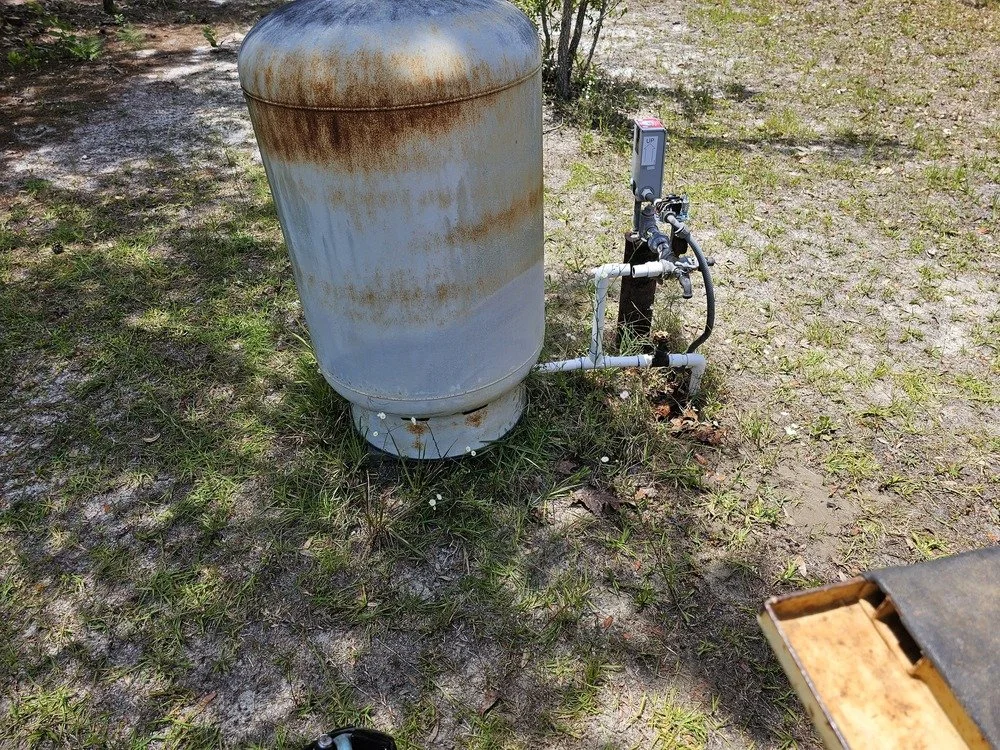 An outdoor water pressure tank with pipes and a digital gauge, situated on grass and dirt ground.