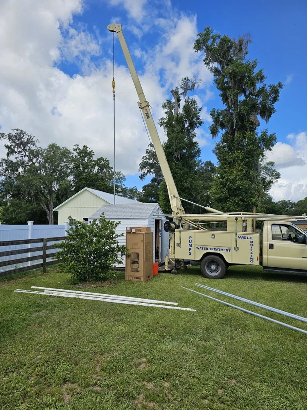 A well drilling truck set up in a backyard with a white house, a wooden fence, and tall trees, under a partly cloudy sky.