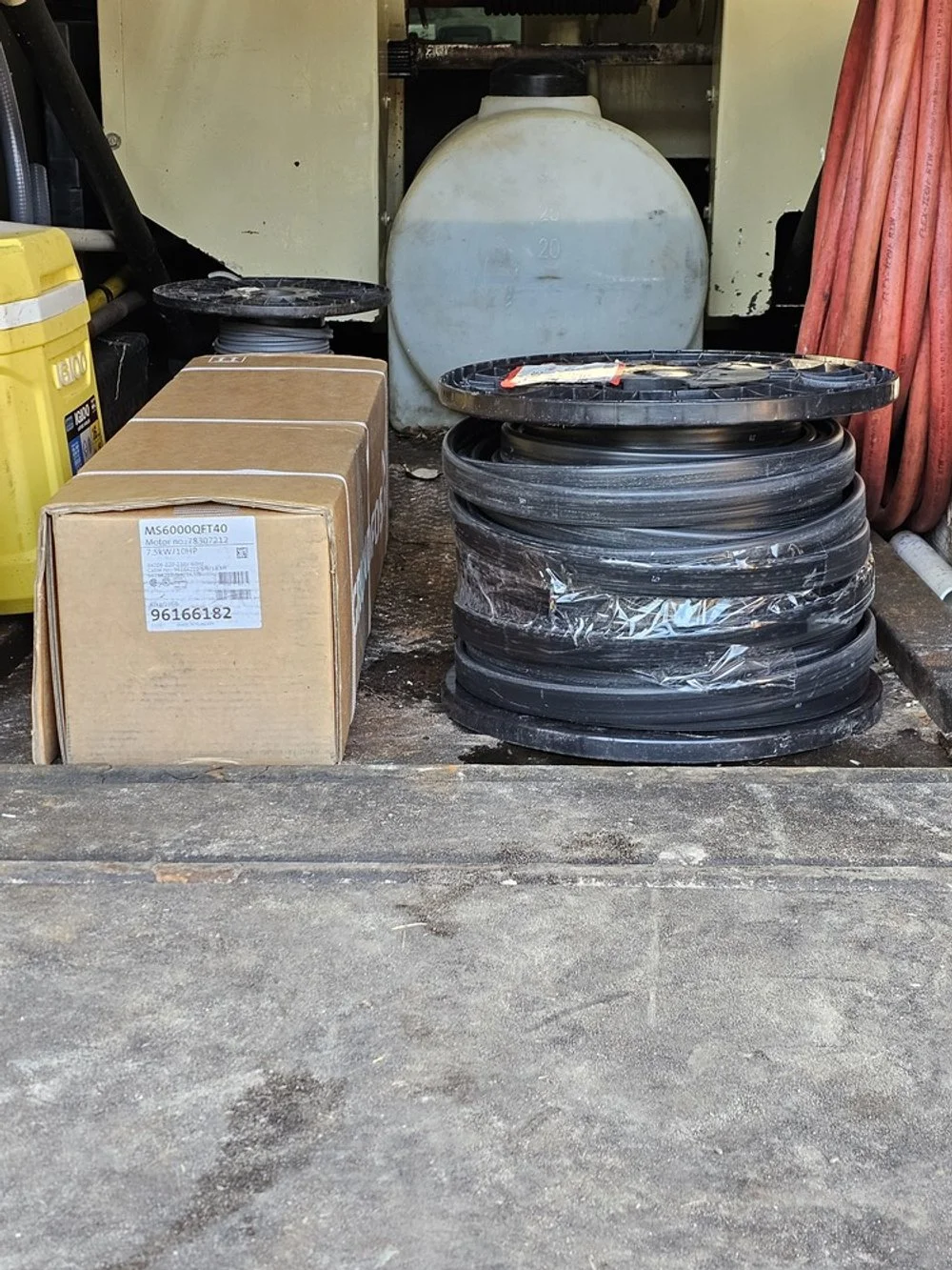 Stacks of black coiled hoses, cardboard boxes, a large white water tank, and orange Extension cords in a workshop or garage.