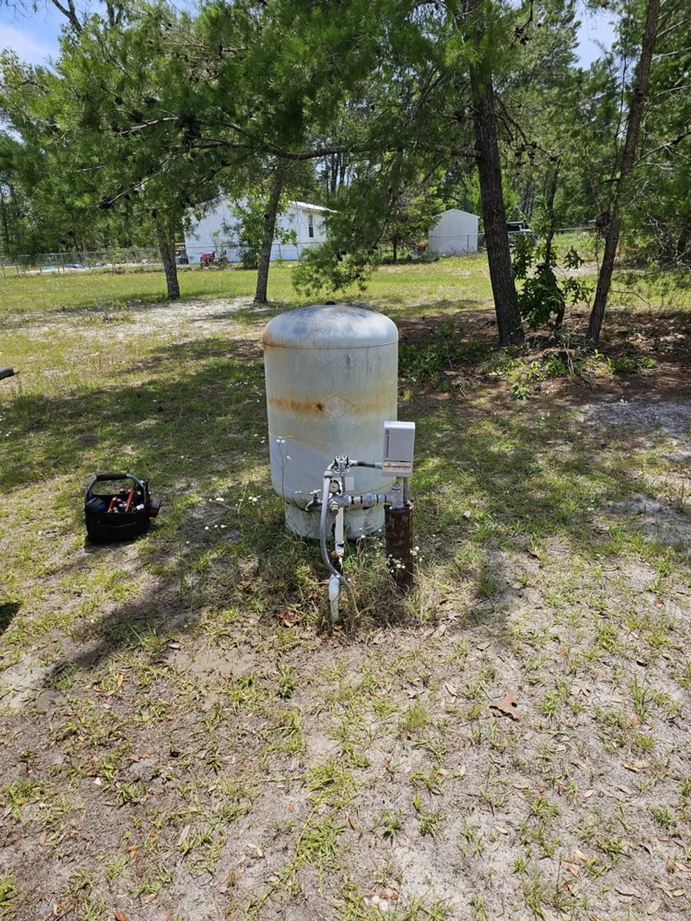 A rusty, white propane tank in a grassy yard with trees and a white house in the background. A black toolbox with tools is placed nearby.