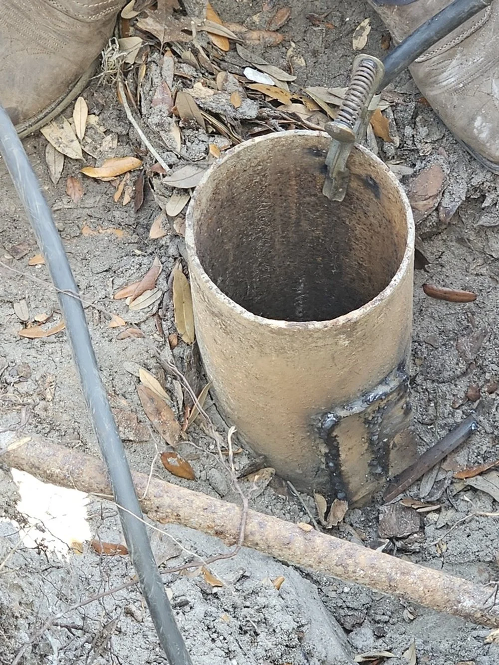 A hole in the ground with a pipe inserted into it, surrounded by dirt and fallen leaves, and a metal tool resting on the hole.