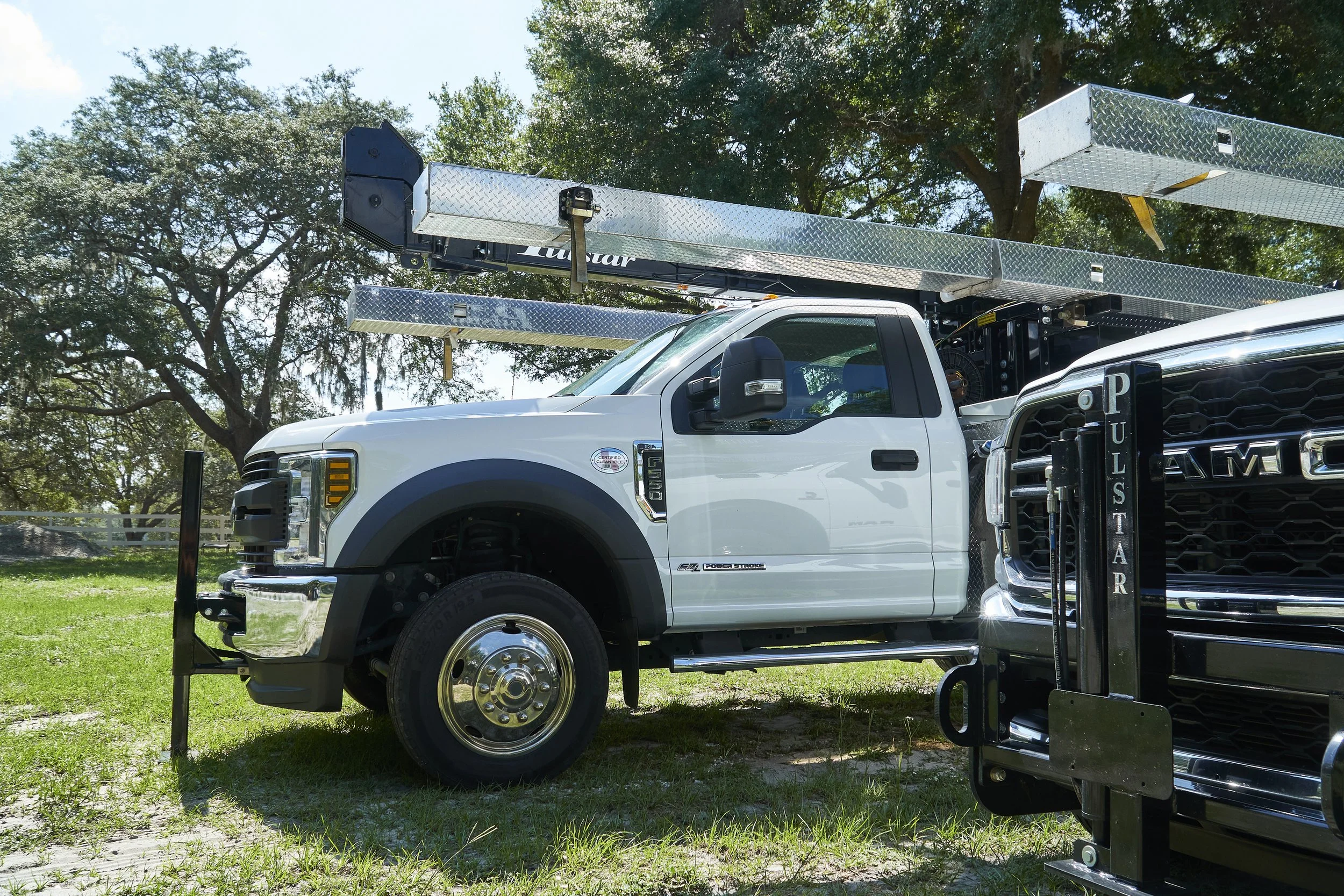 A white utility truck with an extendable lift and long metal shelving, parked on grass with trees in the background.