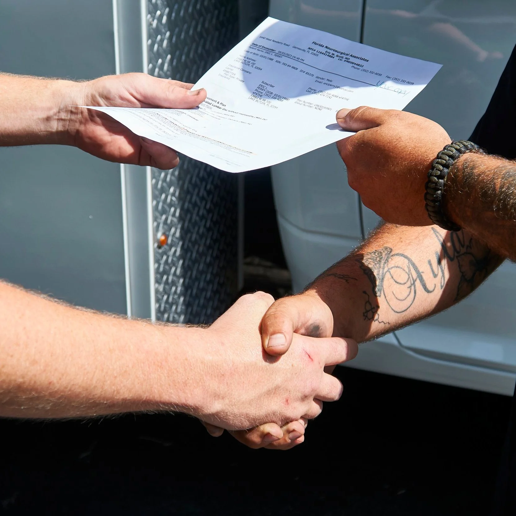 Two people are shaking hands outdoors. One person is holding a medical document, and the other grips the handshake firmly. The person holding the document has tattooed arms and wearing a bracelet.