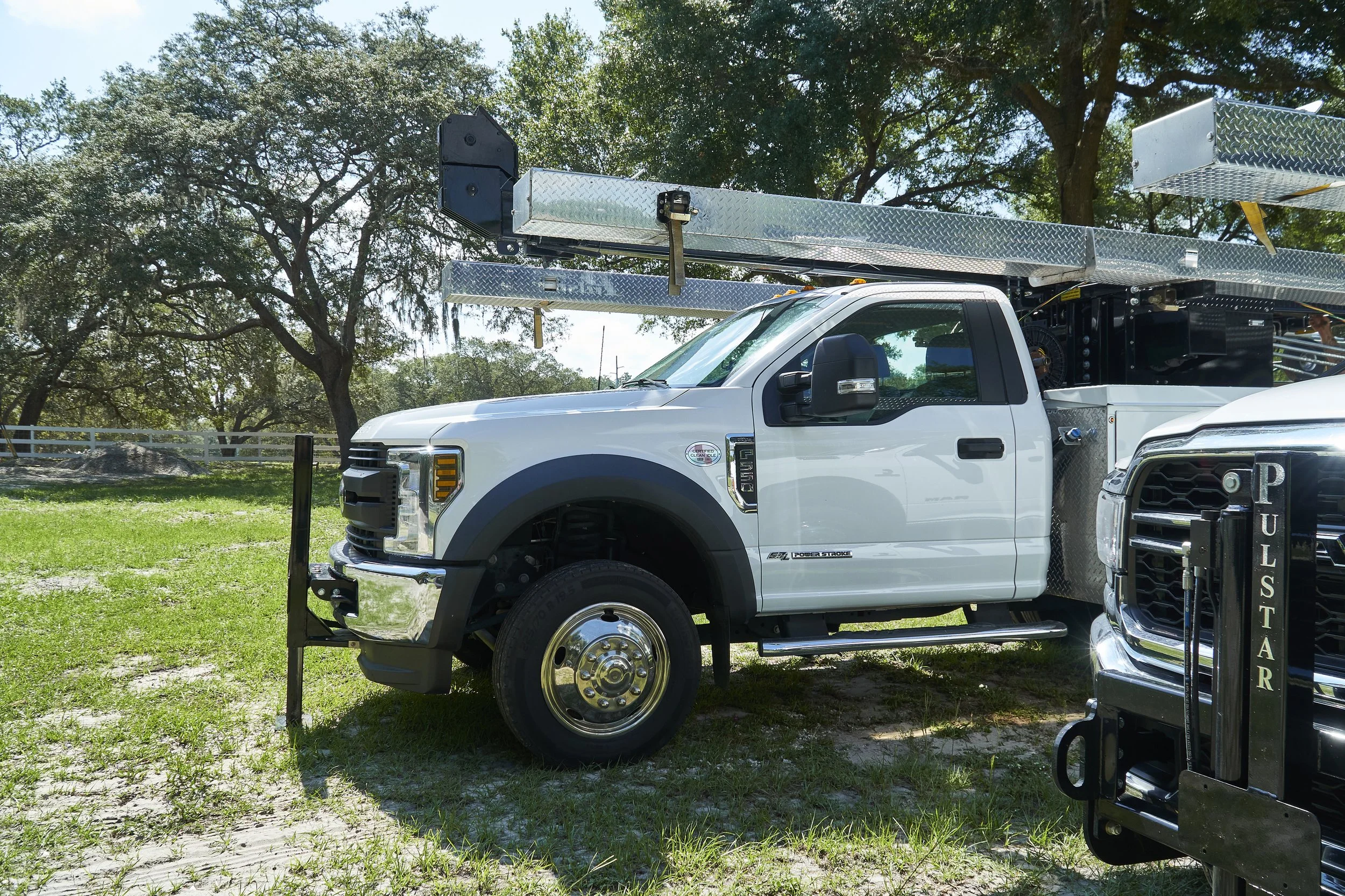 A white utility truck with an extendable boom arm parked on green grass under trees, with a black forklift visible in the foreground.