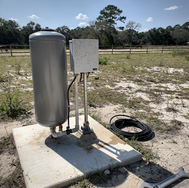 Metal tank and control box with hose on concrete pad in grassy field with fence and trees.